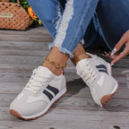 Angled view of grey suede-panel trainers with navy stripes and a white cushioned sole, shown on a wooden deck.