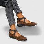 Brown suede ballet flats with elastic crossover straps, worn with cropped grey jeans, side profile
