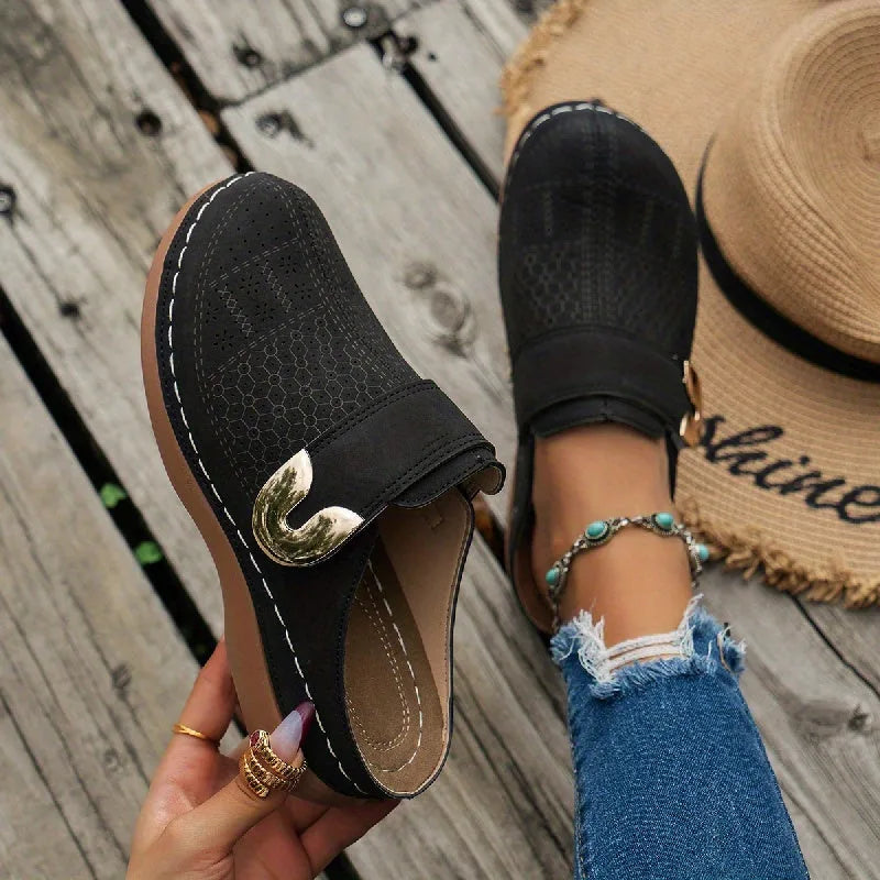 Black suede clog mule with laser-cut floral pattern and gold buckle detail, held by hand beside a model's foot in a straw hat scene
