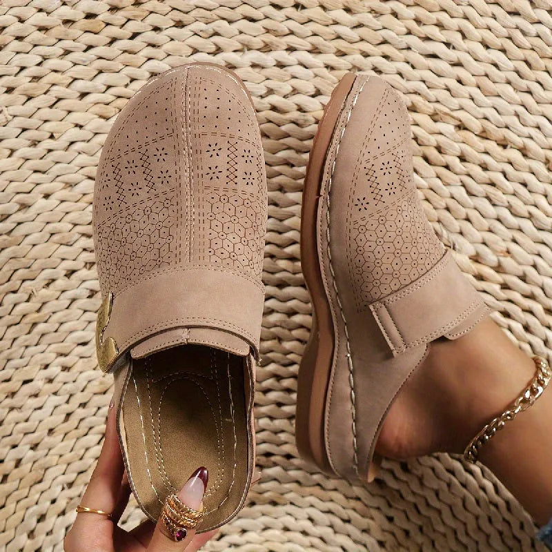 Close-up pair of beige perforated suede clogs with gold hardware and visible cream topstitching on a woven jute mat