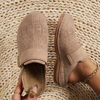 Close-up pair of beige perforated suede clogs with gold hardware and visible cream topstitching on a woven jute mat