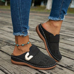 Black suede slip-on mule with honeycomb cut-out pattern worn outdoors on a wooden deck with cropped denim and an anklet