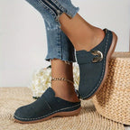 Navy blue suede backless mule with gold buckle accent shown on foot beside woven basket and white flowers