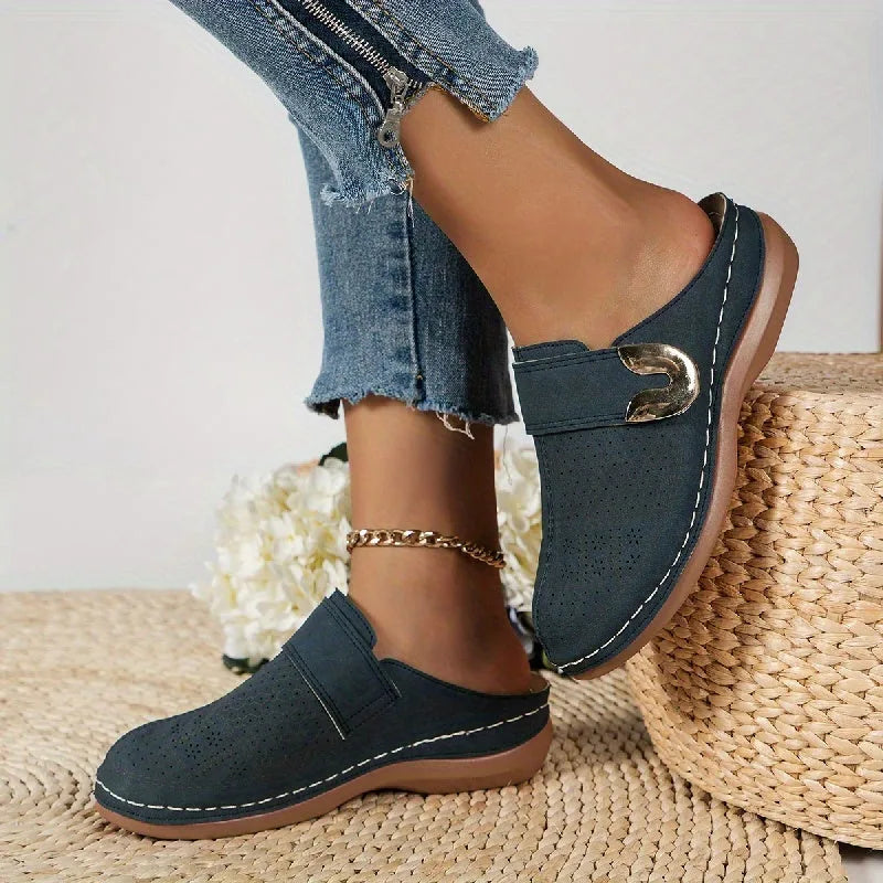 Navy blue suede backless mule with gold buckle accent shown on foot beside woven basket and white flowers