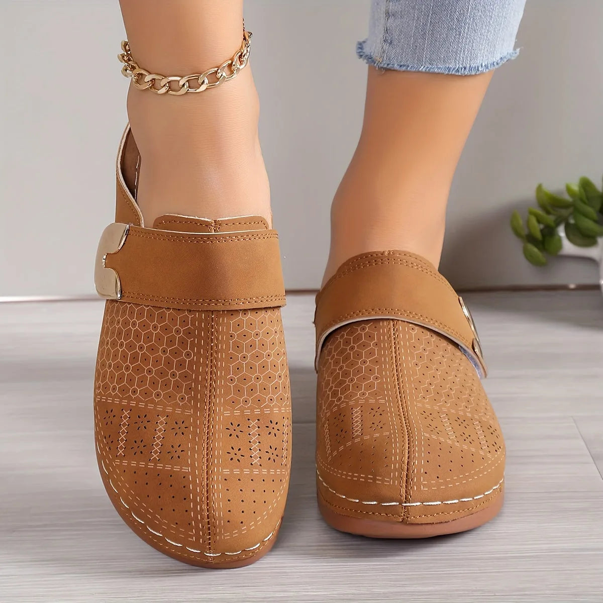 Pair of tan suede clogs with perforated detailing and gold trim displayed front-on against a wood floor backdrop