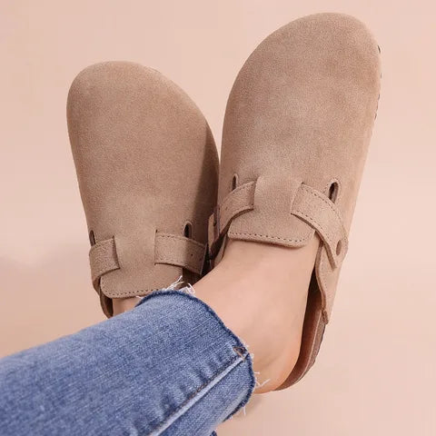 Close-up of taupe suede clog slippers worn on bare feet, showing buckle strap and curved toe shape