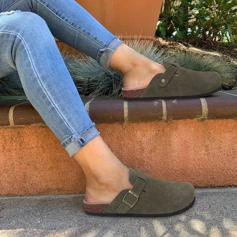 Olive green suede clog slippers worn outdoors with jeans, showing cork-effect sole and buckle detail