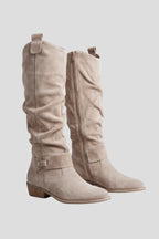 Pair of beige suede knee-high boots with slouchy shaft, back pull tab and ankle strap, shown at an angle