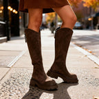 Wide shot of brown suede knee-high boots worn with a pleated skirt, standing on an autumn city pavement