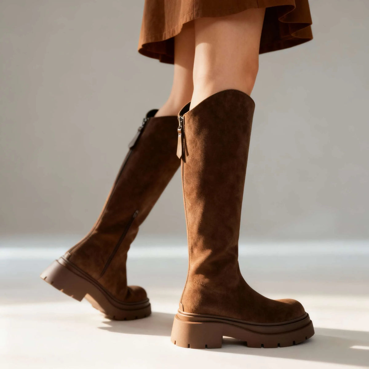 Studio shot of brown suede knee-high boots from behind, showing the back zip pull and heel construction