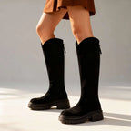 Black suede knee-high boots in profile against a plain studio backdrop, showing smooth shaft and chunky sole