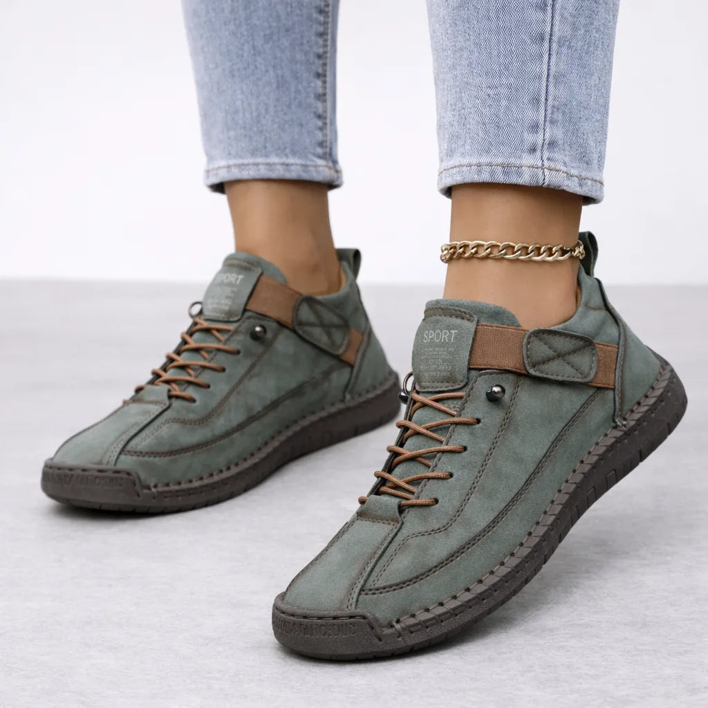 Sage green suede trainers with tan trim detailing and a dark brown stitched outsole, shown from the side on feet