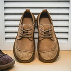 Front view of tan suede derby shoes with contrast lacing, standing side by side against a slatted white panel