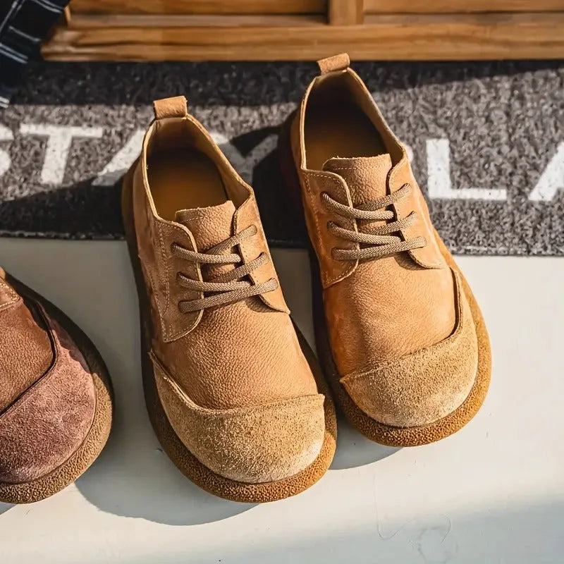 Overhead view of tan suede lace-up shoes on a white floor beside a doormat, showing the split leather and suede toe panel