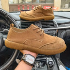 Side profile of a tan leather derby shoe with suede toe cap held above a matching pair on a car dashboard