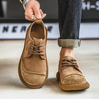 Close-up of a hand holding a tan suede derby shoe by its heel loop, worn with rolled dark denim jeans