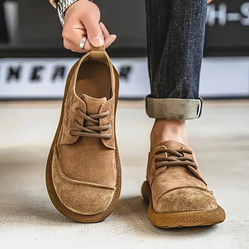 Close-up of a hand holding a tan suede derby shoe by its heel loop, worn with rolled dark denim jeans