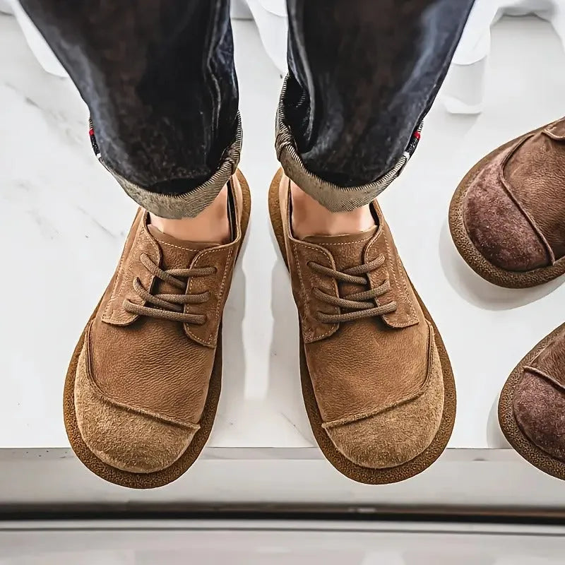 Top-down view of a person wearing tan suede lace-up shoes with turned-up denim trousers on a tiled floor