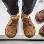 Top-down view of a person wearing tan suede lace-up shoes with turned-up denim trousers on a tiled floor