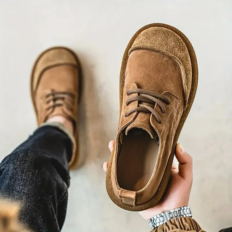 Close-up of a hand lifting a tan suede shoe by the back tab, showing the ridged rubber outsole texture