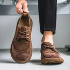 Close-up of a tan suede derby shoe held in hand, showing the beige lacing and stitched seam detail