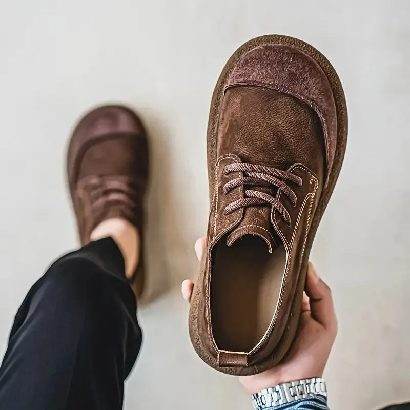 Close-up of a hand holding a dark brown suede lace-up shoe next to a person wearing the matching pair