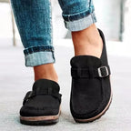 Black suede slip-on clogs with adjustable buckle detail and ridged cork-style sole shown on bare feet with denim.