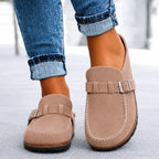 Taupe suede slip-on clogs with buckle trim and stacked sole, close-up front view with cuffed jeans.