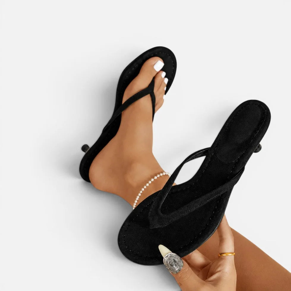 Black suede thong sandals with kitten heel worn on a model's feet, close-up showing toe strap detail