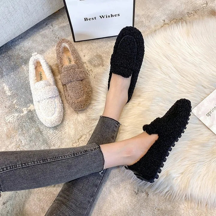 Overhead view of black and cream teddy fleece loafers worn with grey jeans on a fluffy rug beside a gift bag