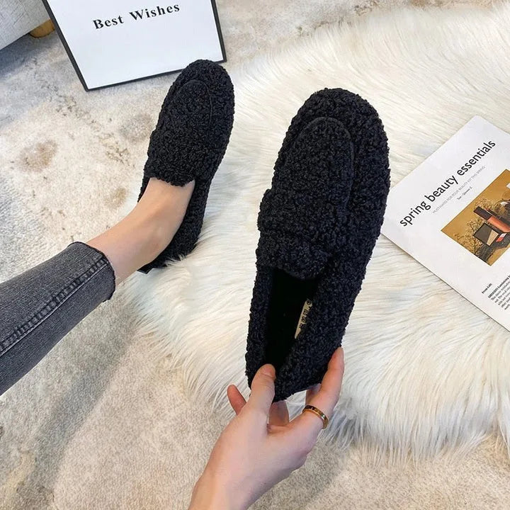 Overhead shot of black teddy fleece loafers held by hand on a cream faux-fur rug