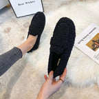 Overhead shot of black teddy fleece loafers held by hand on a cream faux-fur rug