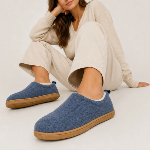 Woman relaxing on floor wearing blue felt slip-on slippers with tan soles, paired with cream loungewear