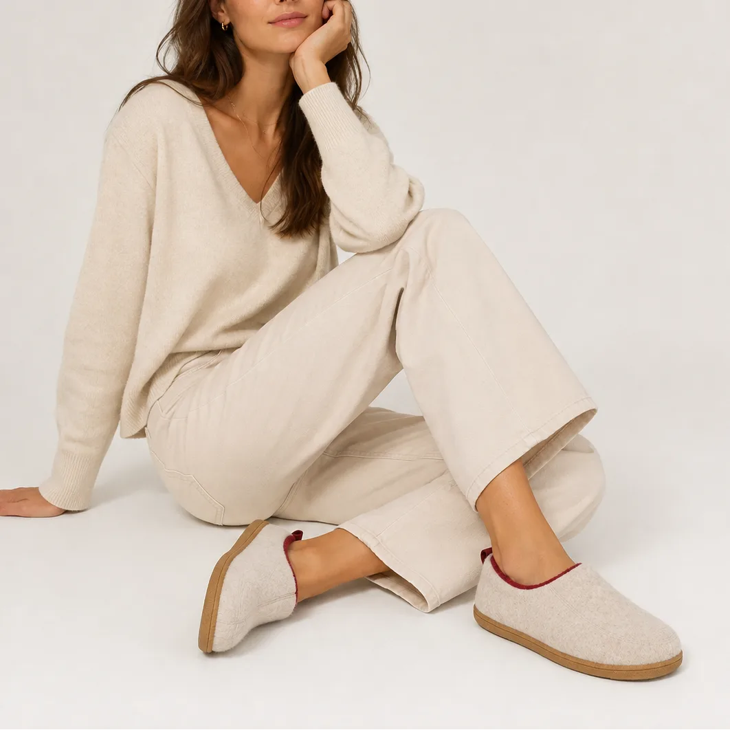 Woman seated wearing beige felt slippers with burgundy trim, styled with a cream jumper and trousers