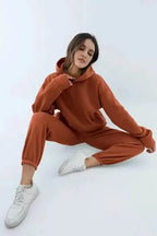 Woman seated wearing a rust orange oversized hoodie and joggers with cuffed hems and white trainers