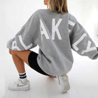 Back view of a mid grey oversized jumper with white letter graphic, paired with white trainers