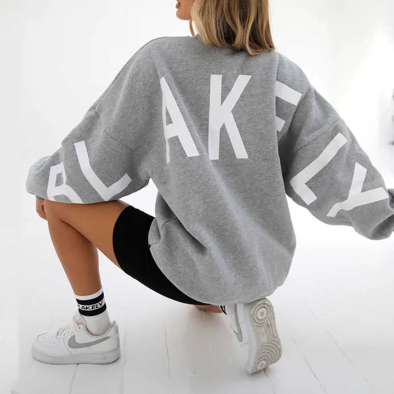 Back view of a mid grey oversized jumper with white letter graphic, paired with white trainers
