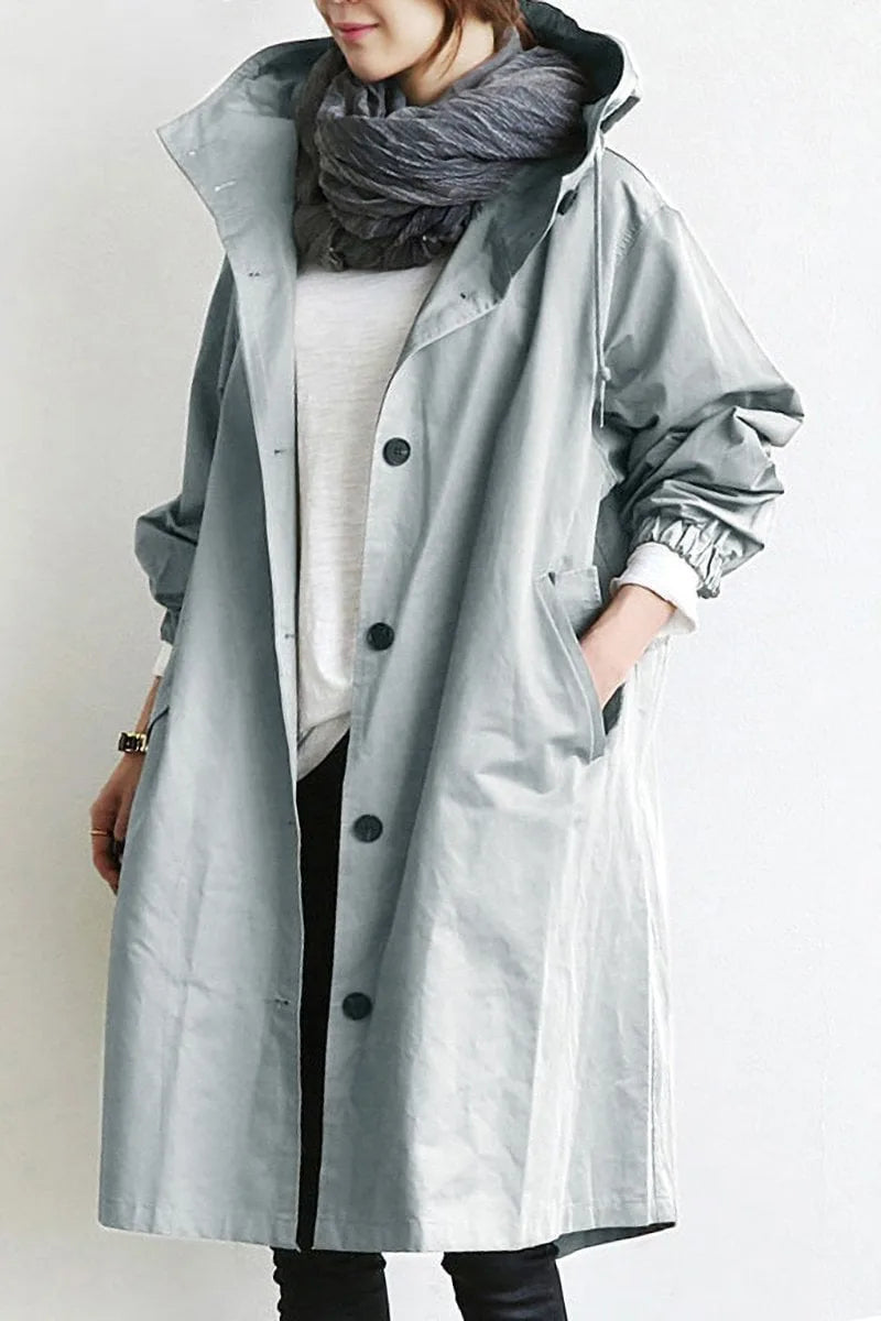 Woman wearing an oversized grey hooded trench coat, unbuttoned over a white jumper and layered scarf