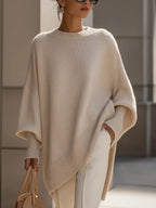 Woman wearing an oversized beige knit poncho jumper with wide neckline, styled with cream trousers and a tan handbag