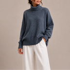Close-up front view of a slate blue oversized polo neck jumper with ribbed collar, worn over white wide-leg trousers.