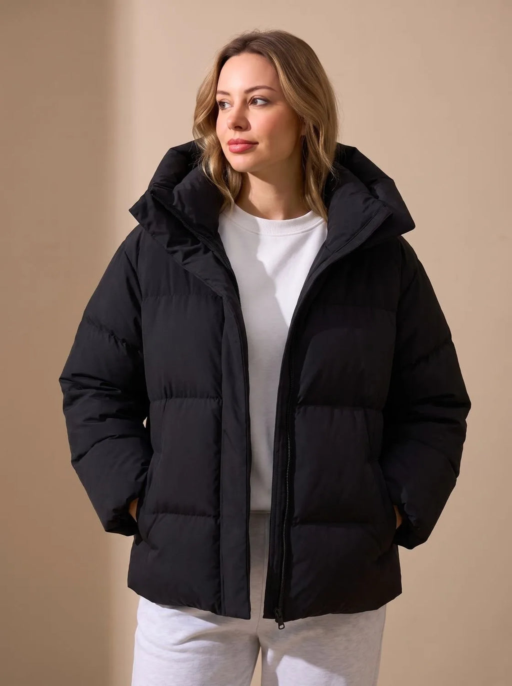 Front view of a black oversized puffer jacket with a padded hood, hands in pockets, worn over a white jumper.