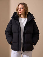 Front view of a black oversized puffer jacket with a padded hood, hands in pockets, worn over a white jumper.