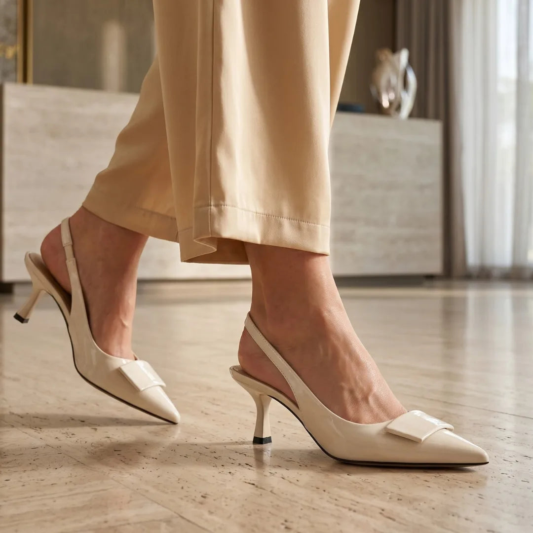 Close-up of cream patent slingback heels worn with beige trousers, shown mid-stride on a warm-toned floor