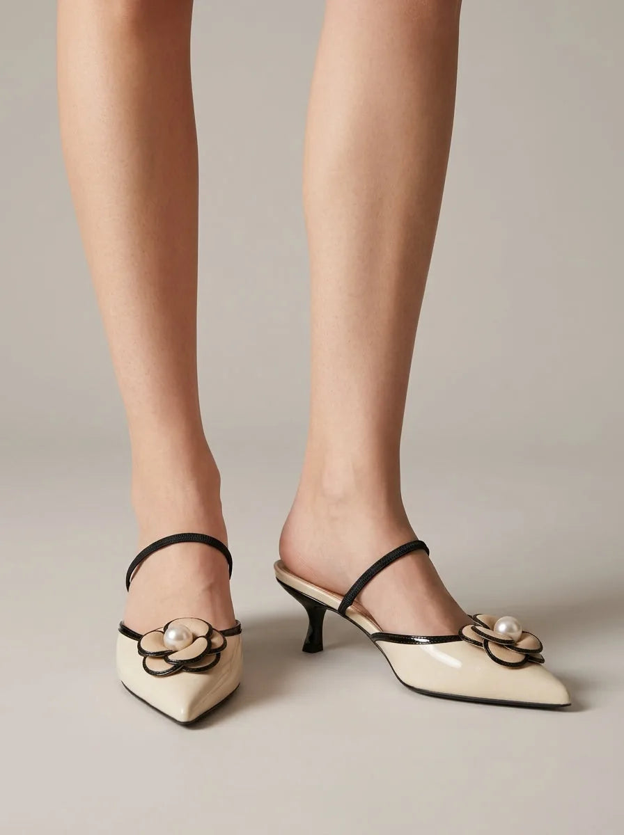 Front view of cream patent pointed-toe mules with slim black strap and pearl floral appliqué at the toe, worn on bare legs