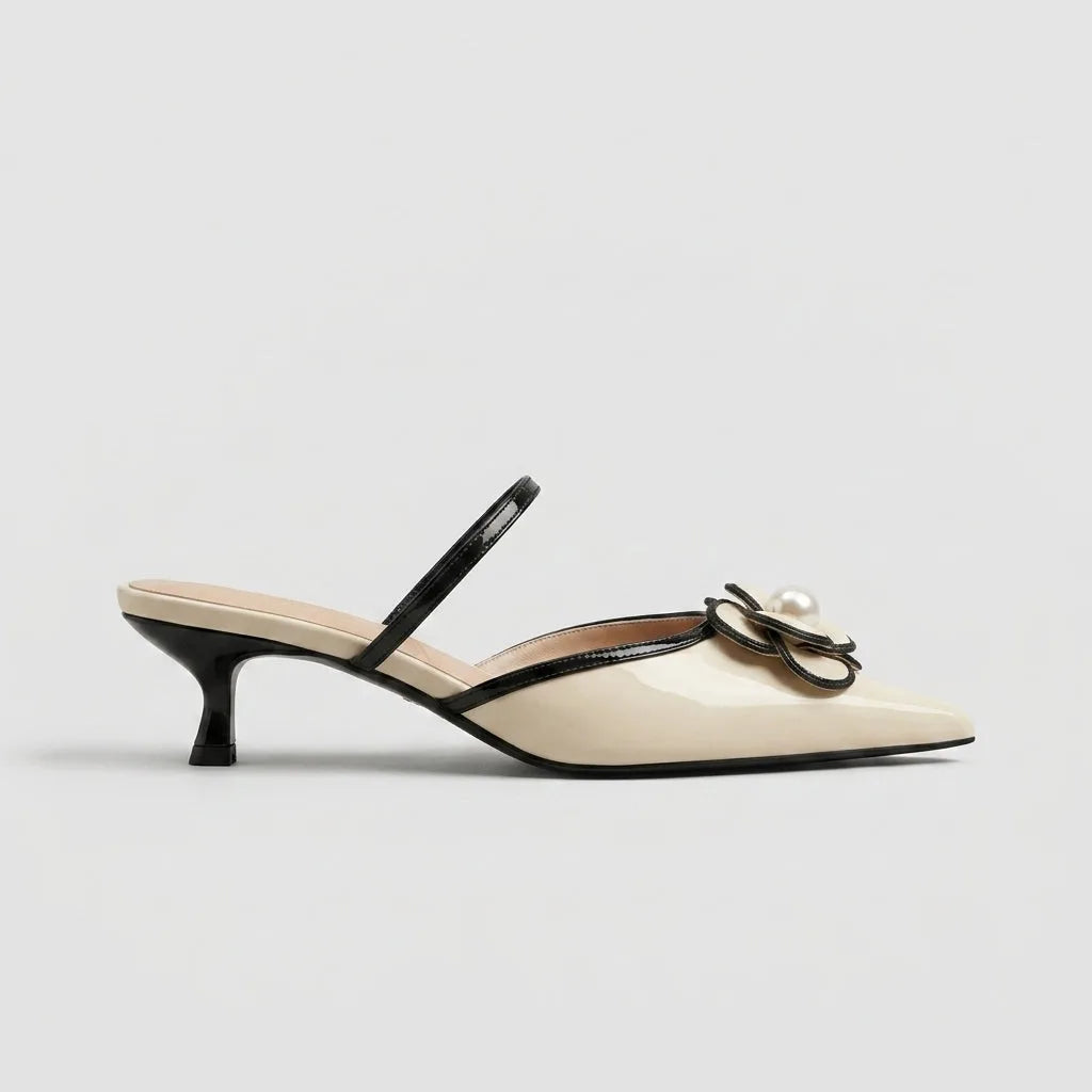 Studio side profile of a cream patent mule with pointed toe, black trim, slingback strap and pearl flower detail on a low kitten heel