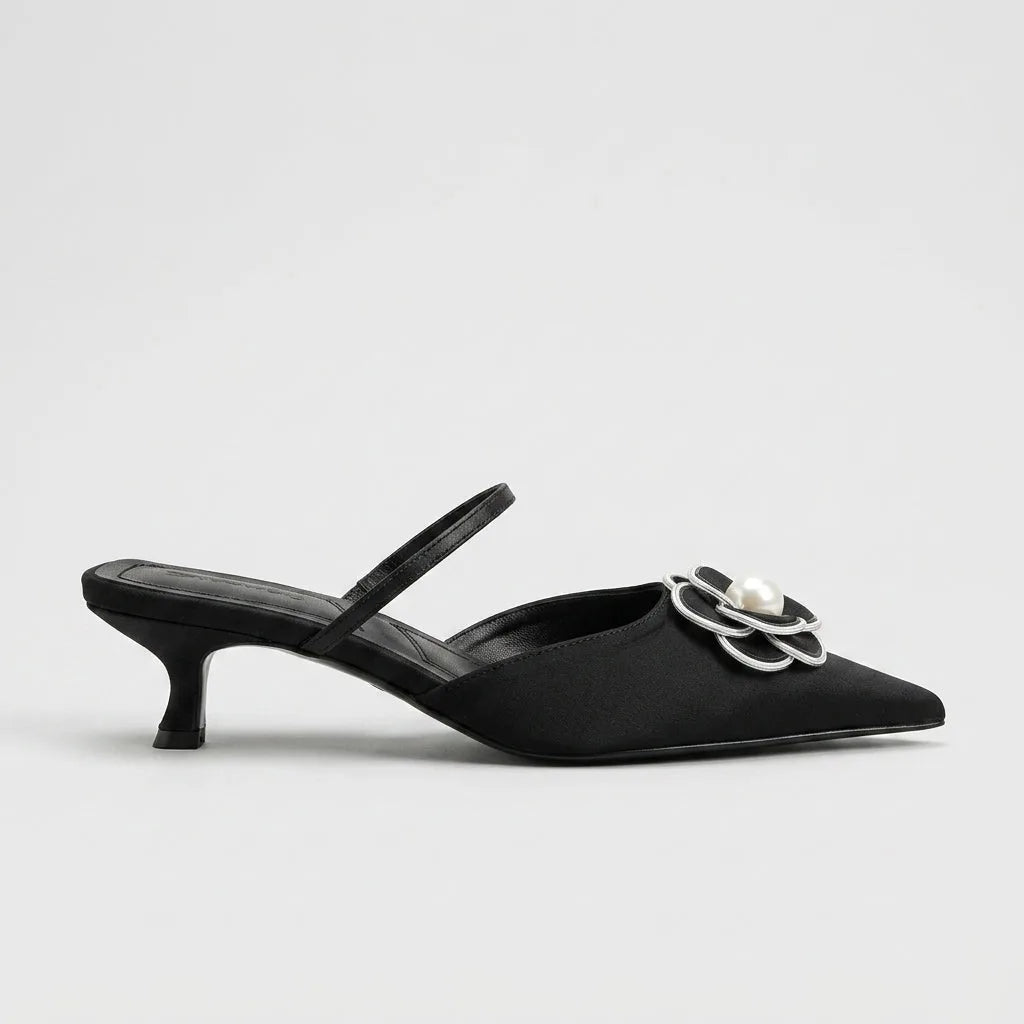 Studio side profile of a black satin mule with pointed toe, slim slingback strap and silver-outlined pearl flower embellishment