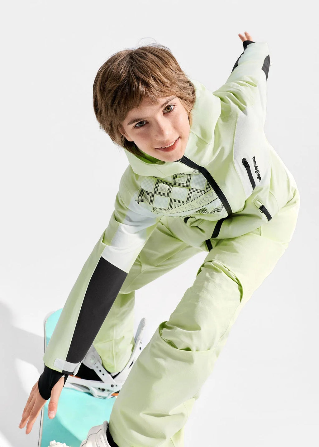 Overhead angle of a boy in a pale green ski jacket bending forward, highlighting the graphic chest panel and taped seams