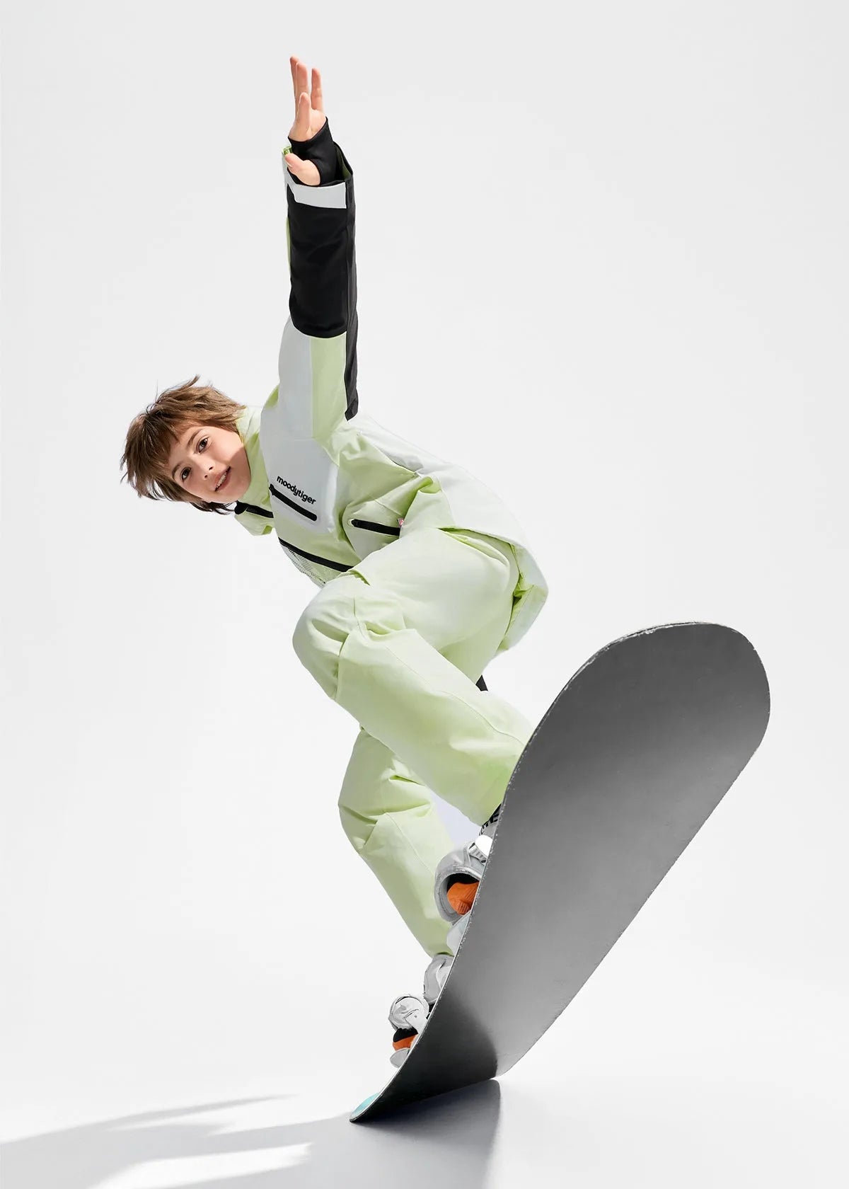 Boy in pale green ski jacket and trousers riding a snowboard, mid-motion with arm raised for balance