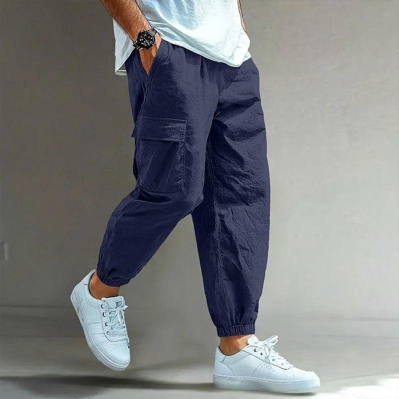 Side view of navy blue cotton cargo trousers with elastic waist, side pocket flap and elastic cuffs, worn with white trainers