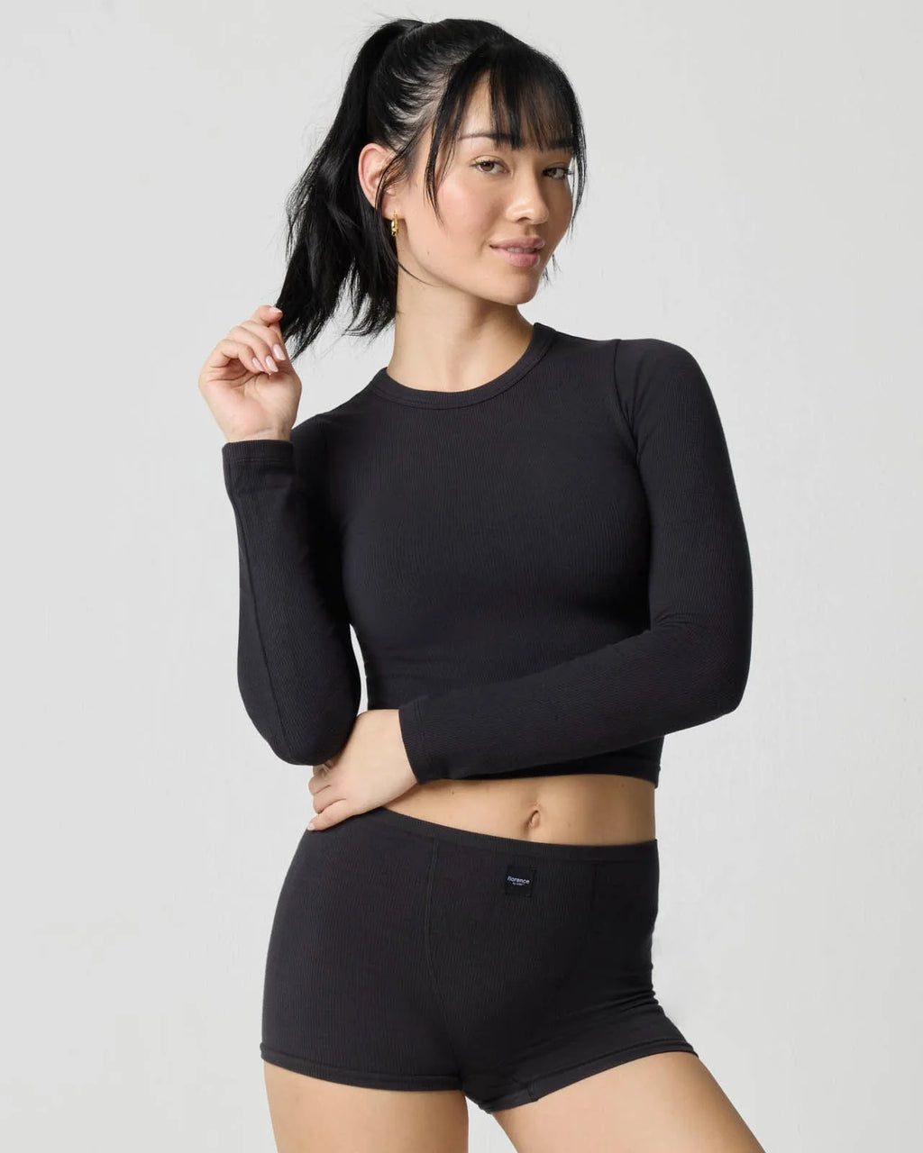 Woman wearing a black ribbed long sleeve crop top with matching shorts, front view with hand touching ponytail